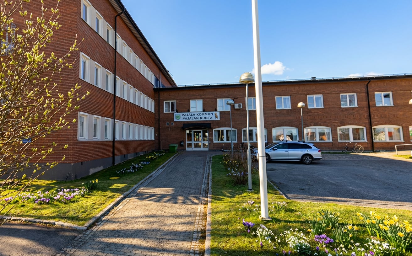 En stor, röd tegelbyggnad med många fönster och en skylt där det står "PAJALA KOMMUN PAJALAN KUNTA" över entrén. Framför byggnaden finns en asfalterad yta med en parkerad silverfärgad bil, en gångväg, gräsytor och vårblommor. En buske med nya löv til