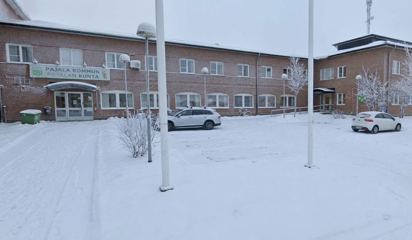 En fasadvy av en tegelbyggnad täckt med snö. Framför byggnaden finns ett snötäckt torg där två bilar, en ljus kombi och en vit halvkombi, står parkerade. Entrén till byggnaden har skyltar med texten "PAJALA KOMMUN" och "PAJALAN KUNTA". Markytan är he