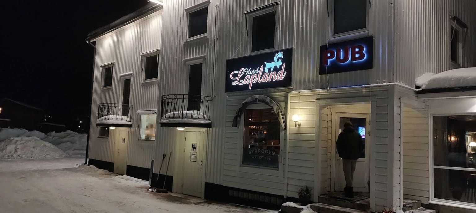 En vit träbyggnad med flera fönster och dörrar under en mörk vinterkväll. Skyltar med texten "Hotell Lapland" och "PUB" är upplysta. Vid en dörr står en person, och snöiga drivor syns runt byggnaden.