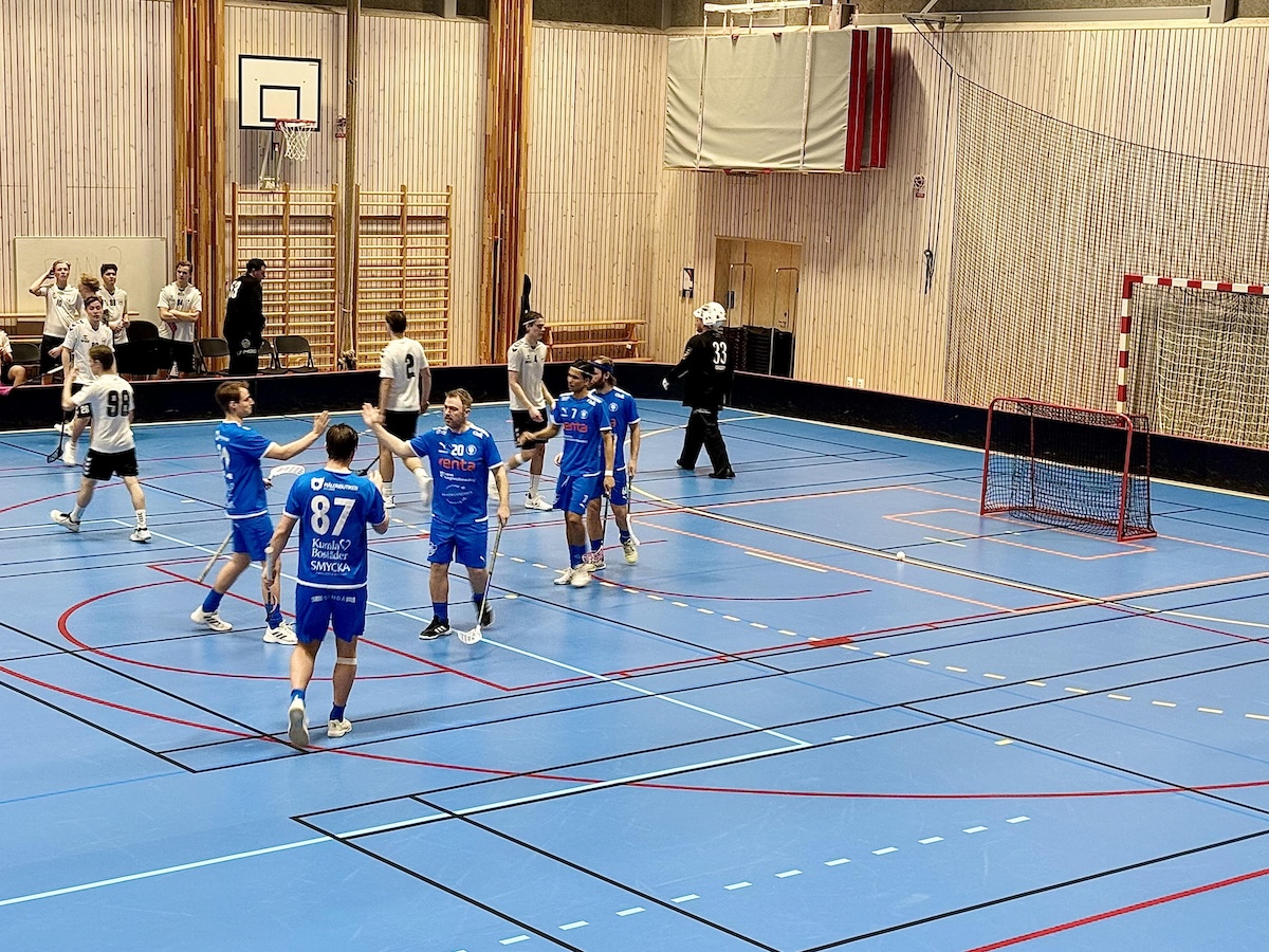 En inomhusbild visar floorballspelare i blåa och vita tröjor på en ljusblå plan med röda, vita och svarta linjer. En spelare i svart målvaktsutrustning syns nära en röd innebandymålbur. Bakgrunden består av ljusa träpanelväggar och ett basketkorg.