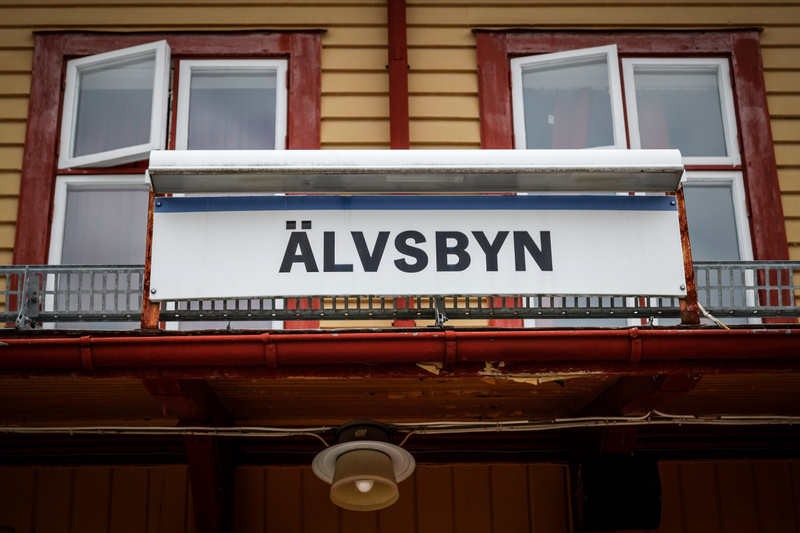 En bild som visar en vit skylt med svart text "ÄLVSBYN", placerad under två fönster med vita karmar på en gul byggnad med röda detaljer. Ovanför skylten finns snö och nedanför sitter en takränna och en lampa.