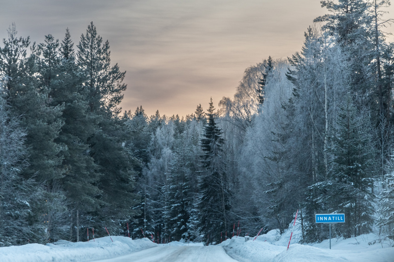 Bilden visar en snötäckt väg som leder in i en skog. På sidorna av vägen finns snövallar och gran- och lövträd som är klädda i snö och frost. En blå skylt med texten "INNATILL" står vid vägkanten. Även röda pinnar är fästa i snödrivorna.