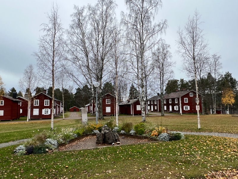 Bilden visar rödmålade trähus med vita knutar och fönsterluckor. Mellan husen och i förgrunden finns gräsmattor och kala lövträd eller björkar. Ett plantering med stenar och olika växter syns i förgrunden. Himlen är grå och molnig.