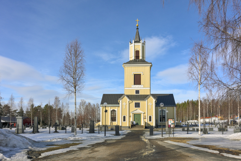 En gul kyrka med ett högt torn och en spira med ett kors på toppen syns centralt i bilden. Runt kyrkan finns en kyrkogård med ett flertal gravstenar och smidesstaket. Delar av marken är snötäckt och några björkar utan löv står runt omkring. Blå himme