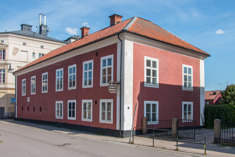En röd byggnad med vita knutar och många fönster står vid en gata under en klarblå himmel. På byggnadens fasad hänger en skylt med texten "ARBOGA MUSEUM". Delar av byggnaden ligger i skugga.