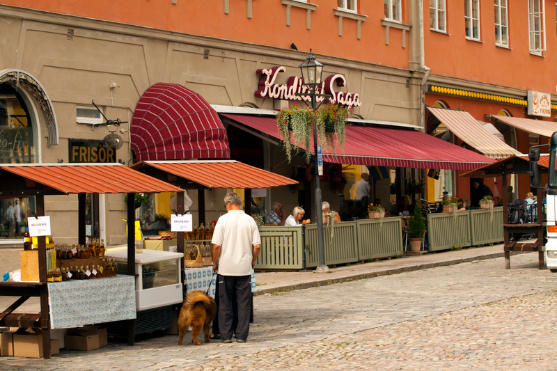 En gatumiljö med en man och en hund framför marknadsstånd. En konditori med uteservering och gäster syns i bakgrunden. Gatan är belagd med kullersten.