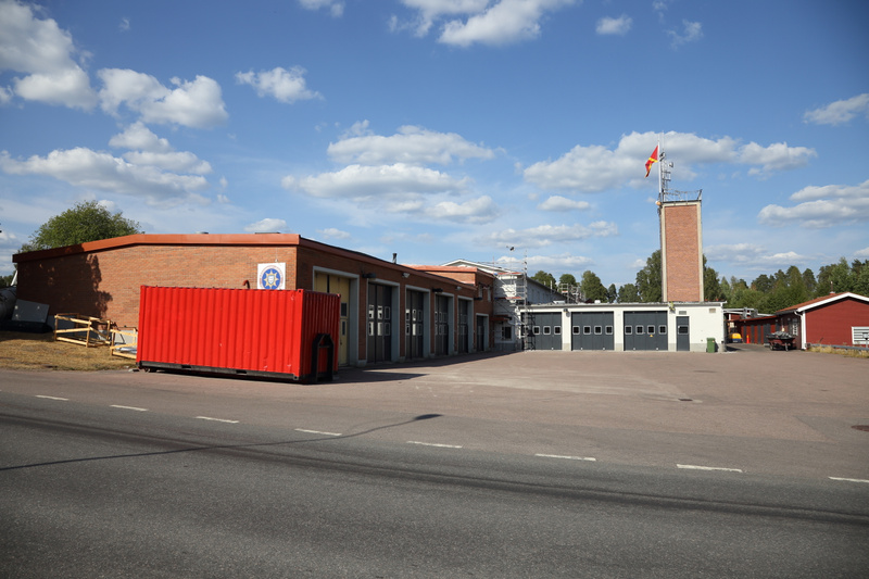En bild visar en tegelbyggnad med många garageportar under en blå himmel med moln. Till vänster står en röd container, och till höger finns ett tegelklätt torn med en röd och gul flagga. En väg löper i förgrunden.