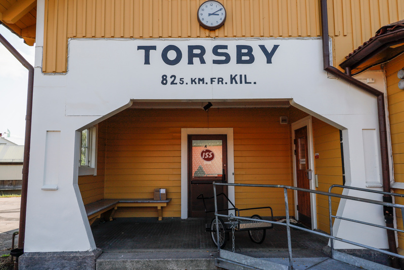 En gul och vit byggnad med texten "TORSBY 825. KM. FR. KIL." ovanför en entré med en brun dörr. En klocka sitter centrerat ovanför texten. Till vänster finns en träbänk med en kartong på, och till höger en liten vagn och en ramp med räcke.