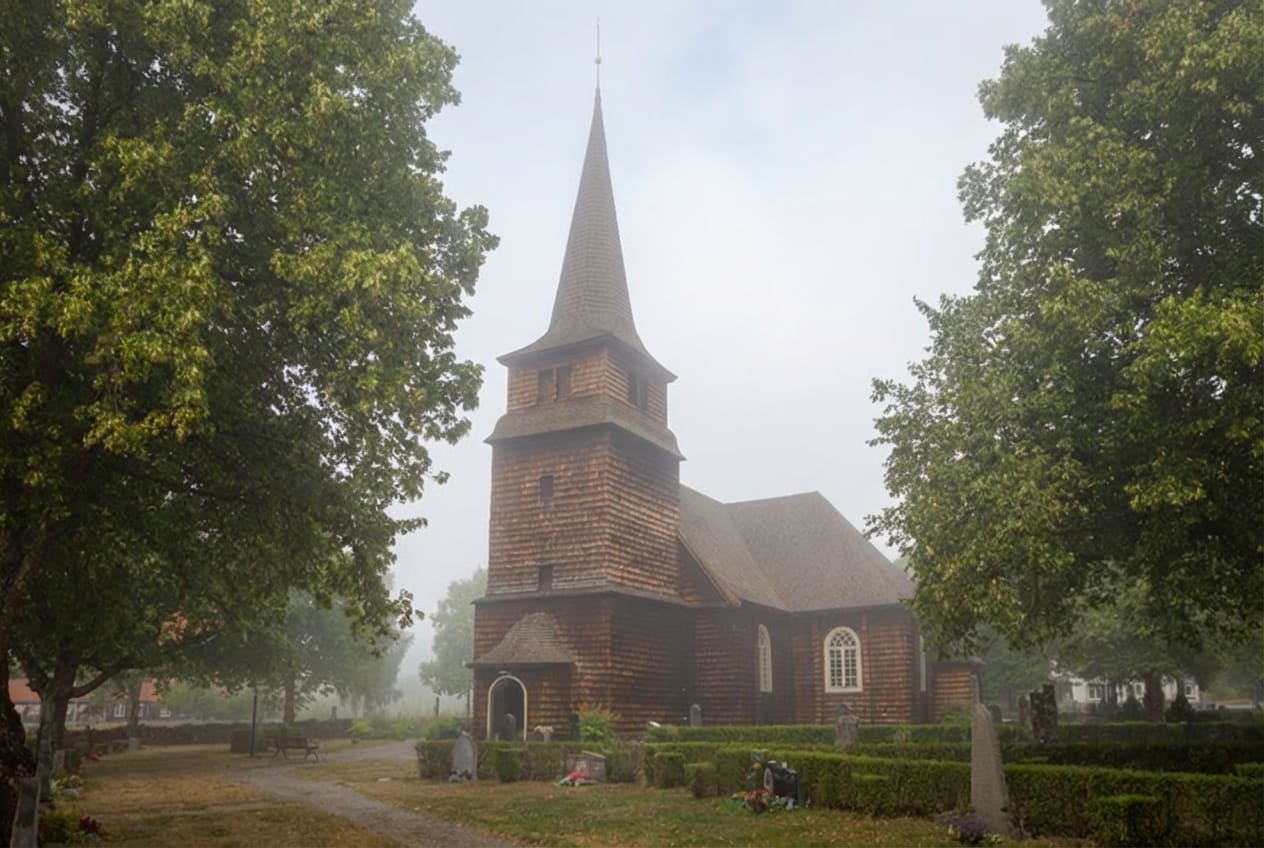 En kyrka med träspånfasad står i dimma, omgiven av träd och gravstenar. Byggnaden har ett spetsigt torn och vitmålade fönster. En grusväg leder till kyrkan, och buskar planterade runt gravstenarna är synliga i förgrunden.