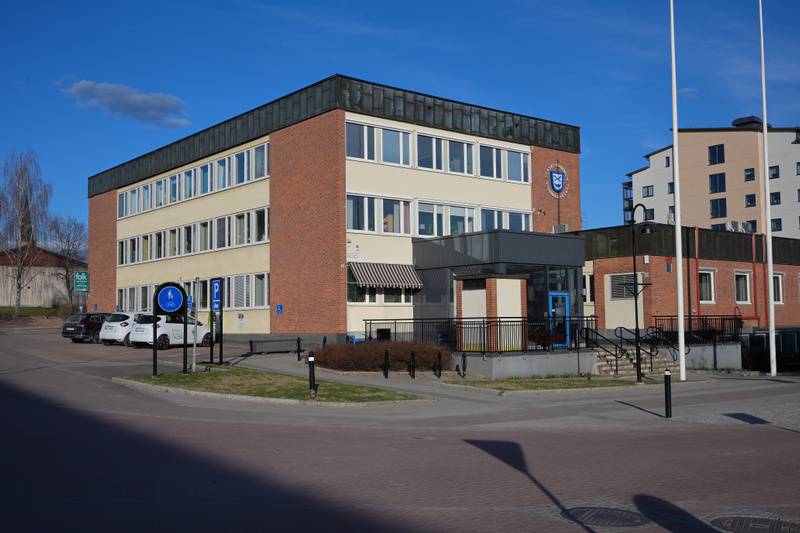 En bild som visar en tegelbyggnad med flera fönster i ett stadsmiljö. Framför byggnaden finns parkerade bilar, blåa skyltar, buskar och en asfalterad yta. Himlen är klarblå och solen lyser, vilket skapar skuggor på marken.