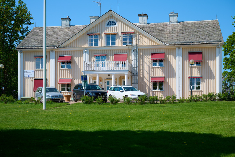 En gul trähusbyggnad med vit trim och röda markiser syns under en blå himmel. Framför byggnaden finns en gräsmatta, buskar och tre personbilar parkerade. En flaggstång står till vänster om byggnaden.