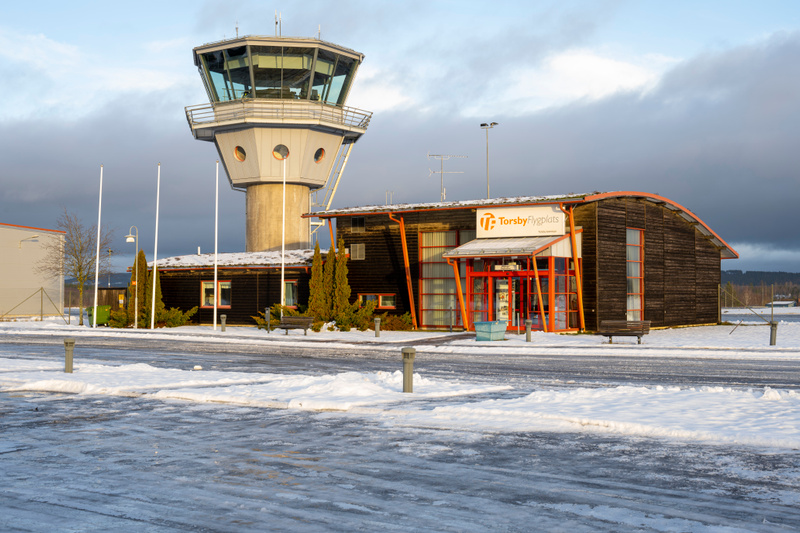 En bild visar en flygplatsbyggnad med ett kontrolltorn bredvid. Framför byggnaderna finns en yta täckt med snö och is. Skylten på byggnaden anger "Torsby Flygplats".