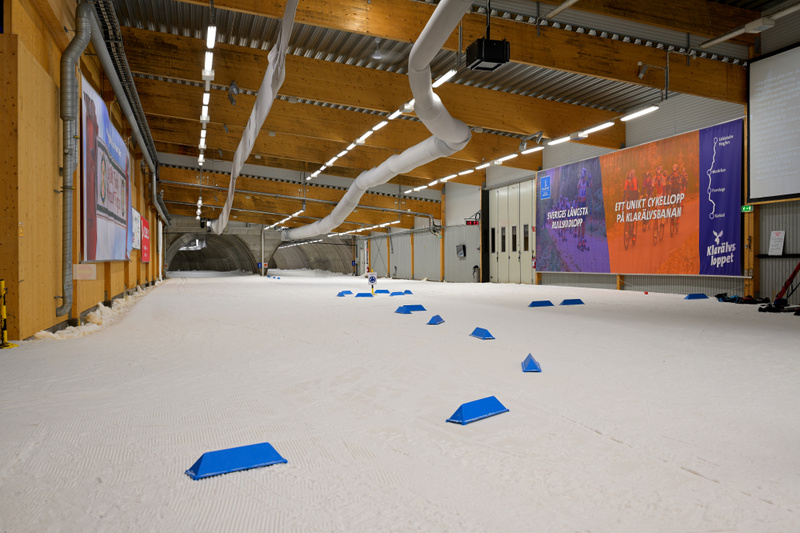 En stor inomhusanläggning med snötäckta markytor, troligen för skidåkning. Taket består av synliga träbalkar och ventilationsrör. Längs väggarna hänger stora banderoller med text och bilder.