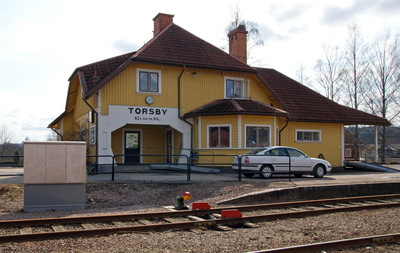 En gul byggnad med rött tegeltak, identifierad som "TORSBY" stationsbyggnad, står bredvid ett järnvägsspår. En ljus bil är parkerad intill byggnaden.
