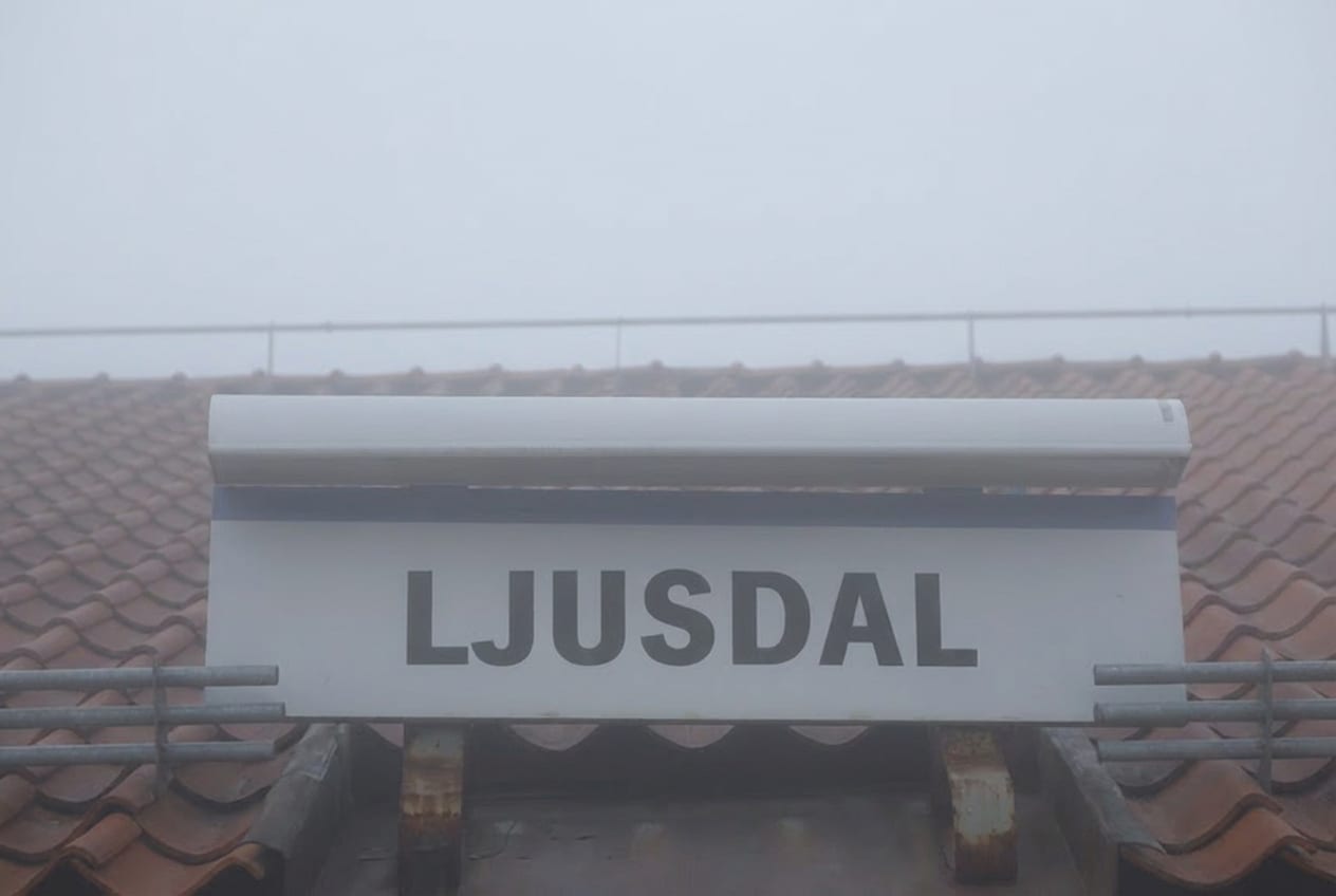 En nära bild av en vit skylt med texten "LJUSDAL" i en svart sans-serif font, delvis skymd av dimma. Skylten är placerad framför ett tak med tegelpannor.