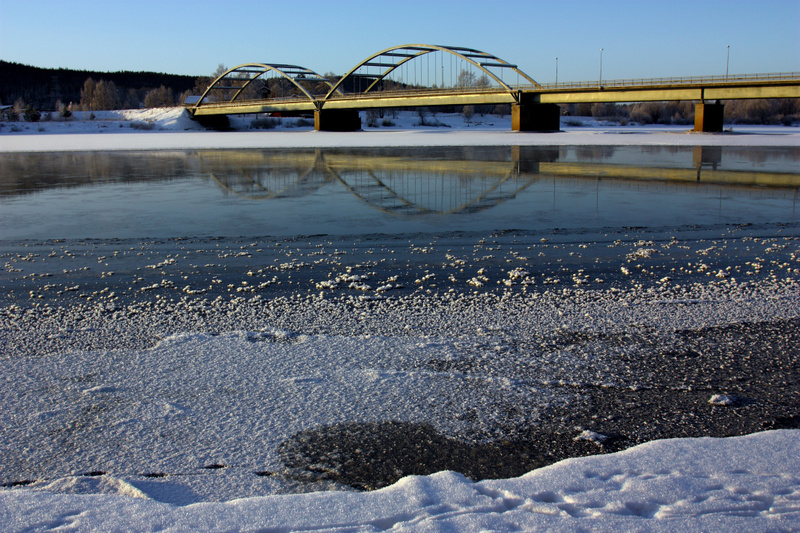 En bro med två valv spänner över en delvis frusen älv eller flod. Vattenytan reflekterar brons valv, och stranden i förgrunden är täckt av snö och is. I bakgrunden syns skogklädda kullar under en klarblå himmel.
