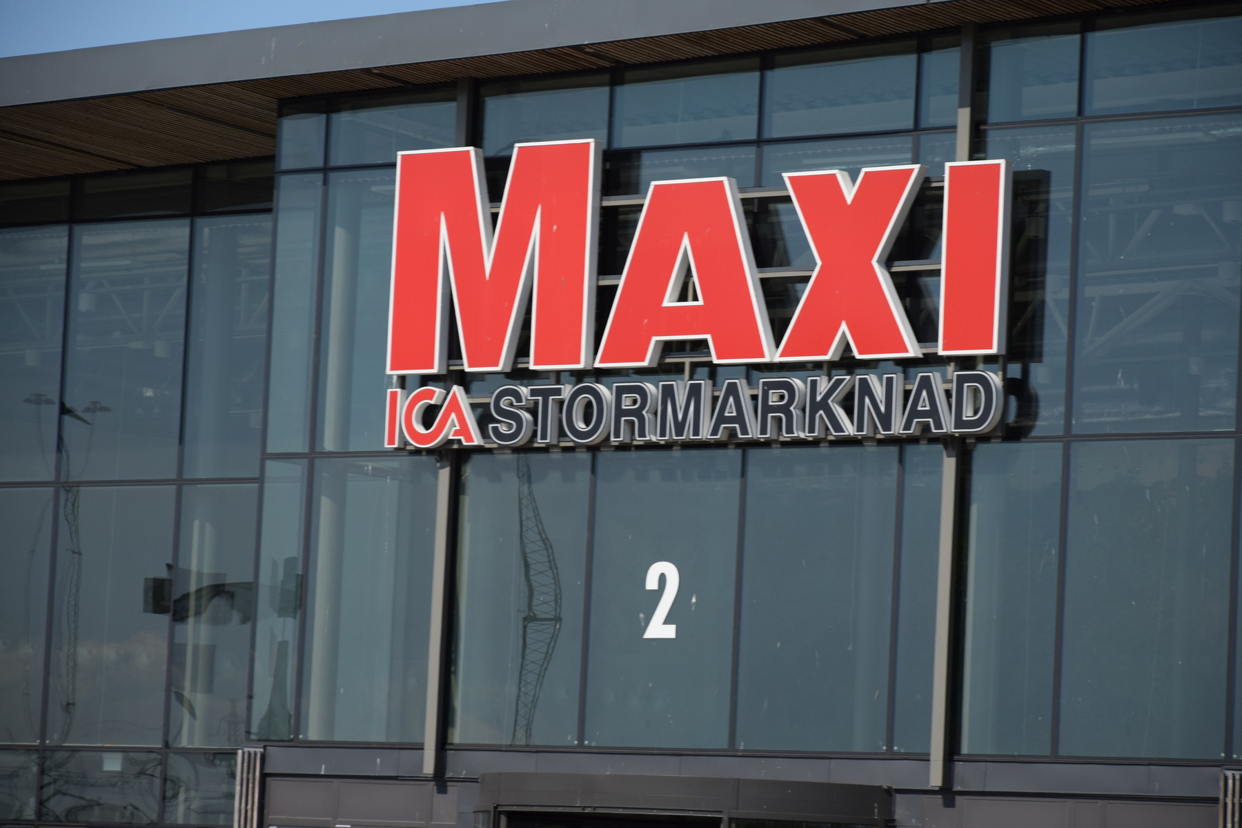 En bild som visar en del av en byggnadsfasad med stora glaspartier. På fasaden sitter en röd skylt med texten "MAXI" i versaler ovanför texten "ICA STORMARKNAD". Siffran "2" syns också på en av glasrutorna.