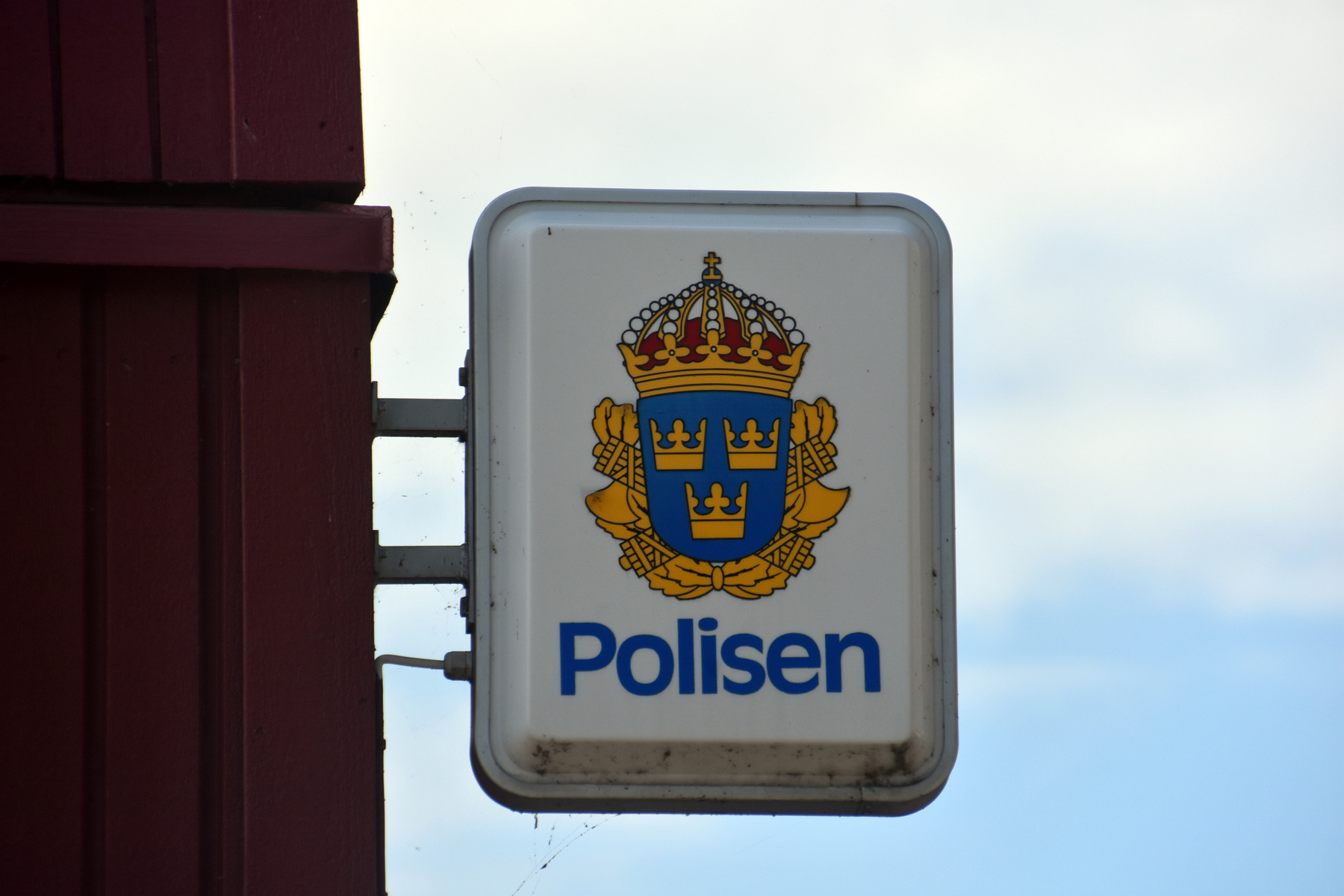 En närbild av en skylt med Polismyndighetens emblem och texten "Polisen". Skylten är monterad på en rödbrun trävägg mot en ljusblå himmel med moln. En del smuts och spindelnät syns på skyltens nedre del.