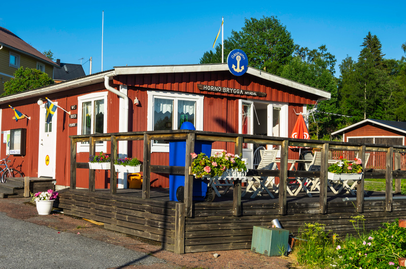En röd stuga med vit knutbräda och en träveranda. På taket syns en blå skylt med ett ankare och texten "Gästhamn". Flera blomlådor med blommor är placerade på verandan. Två svenska flaggor hänger vid ingången. En röd parasoll och möbler finns på vera