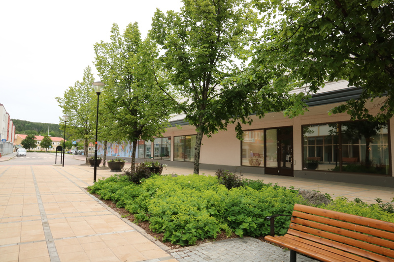 En gata i en stad med en trottoar till vänster, en rad träd och buskar i mitten, och butiksfasader till höger. I bakgrunden syns fler byggnader och en skogsklädd kulle under en molnig himmel. En träbänk är placerad i förgrunden till höger.
