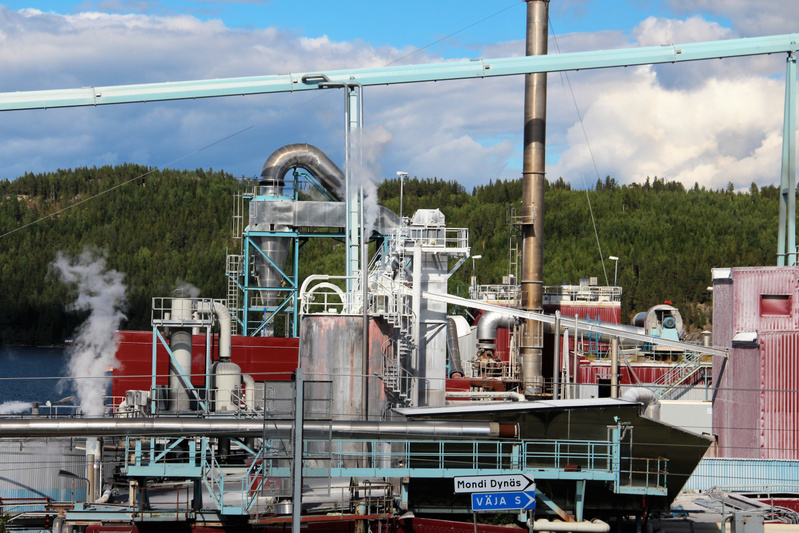 En industriell anläggning med omfattande rörsystem, tankar, skorstenar och olika byggnadsstrukturer. Vit ånga stiger upp från flera punkter. I bakgrunden syns skogklädda kullar och en del av ett vattendrag. En blå vägskylt visar riktning. Himlen är d