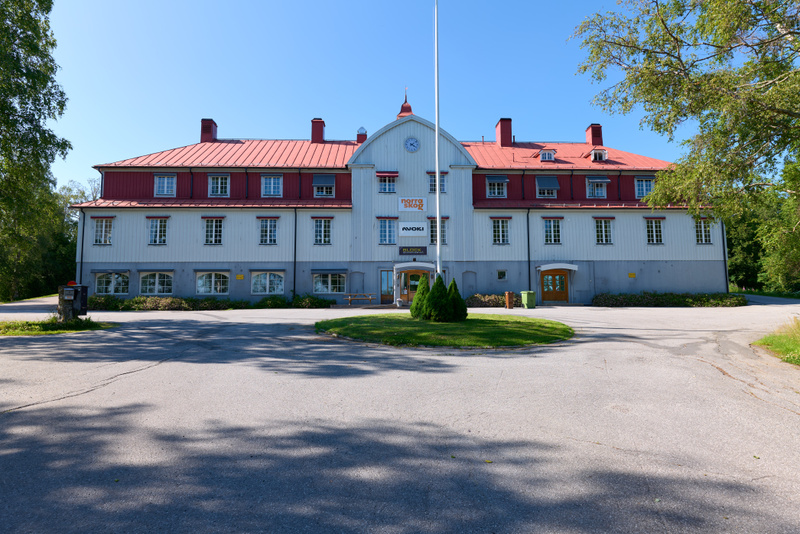 En stor, ljus byggnad med rött tak och vita väggar. Byggnaden har många fönster och en central entré med en flaggstång framför. Framför byggnaden finns en asfalterad yta och träd runtomkring. Himmelen är klarblå.