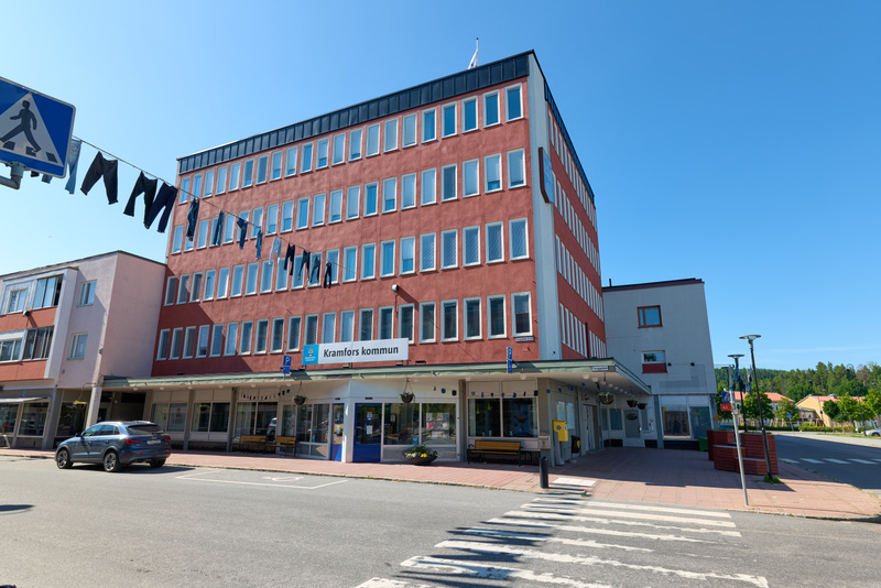 En röd byggnad med många fönster dominerar bilden under en klarblå himmel. Framför byggnaden finns en skylt med texten "Kramfors kommun" och flera kläder hänger på en lina. En personbil är parkerad till vänster och ett övergångsställe syns i förgrund