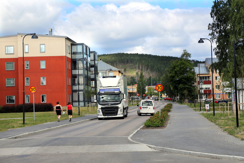 En gata med flerbostadshus på vänster sida och berg i bakgrunden. En vit lastbil, en vit skåpbil och en röd personbil syns på vägen. Två personer går på gångbanan till vänster. En 30 km/h skylt står vid vägkanten. Himlen är delvis molnig.