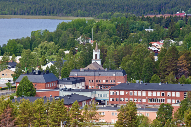 En översiktsbild av en stadsmiljö med en kyrka, flera byggnader och grönskande träd, belägen vid en sjö. Berget i bakgrunden är täckt av barrträd. Flera tegelsbyggnader med mörka tak framträder i förgrunden.