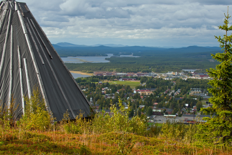 En vidvinkelbild tagen från hög höjd som visar en tältliknande struktur i förgrunden, en stad med bebyggelse och skog i mitten, samt vatten och berg i bakgrunden under en molnig himmel.