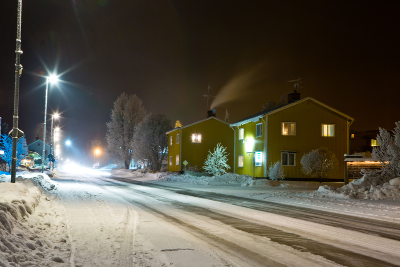 En vinterbild tagen på natten visar en snötäckt gata med två gula hus på höger sida. Gatan är upplyst av gatlyktor och ljuset från husens fönster. Träd och buskar täckta med snö syns längs vägen och vid husen. Från ett av husen stiger rök ur en skors