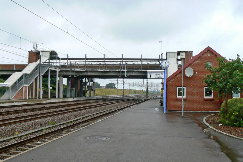 En översiktsbild av en tågstation med spår, perronger och en röd tegelbyggnad. En gångbro och trappor syns i bildens övre vänstra del. En skylt med texten ”Spår 1 Tåg mot Helsingborg och Malmö” samt en klocka är placerade vid gångbron.