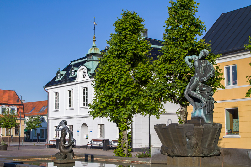En bild av ett torg i dagsljus med flera byggnader och skulpturer vid en fontän. I bakgrunden syns vita och gula byggnader med tak, samt gröna lövträd. En större bronsfärgad skulptur står i förgrunden vid en fontän.