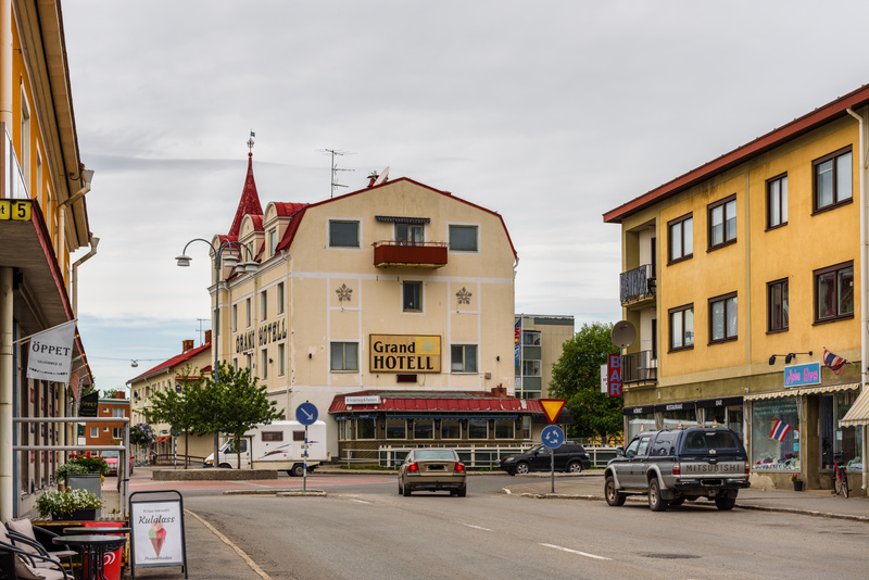 En ganska bred bild av en gata i en stad med byggnader på båda sidor under molnig himmel. Två bilar är parkerade till höger och en rör sig bort från kameran. En husbil syns parkerad till vänster och en skylt som gör reklam för "KULGLASS" står på trot