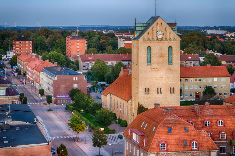 En översiktsbild sedd ovanifrån av en stadskärna med en stor kyrka i förgrunden, omgiven av bostadshus med röda tak och en gata med bilar. I bakgrunden syns skog och hus samt vindkraftverk vid horisonten.