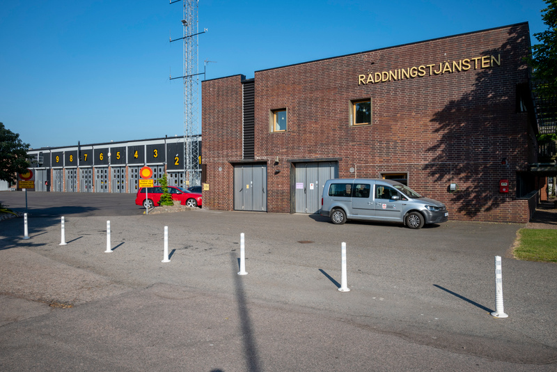En bild som visar en brandstation med flera garageportar numrerade 1-9. I förgrunden syns vita stolpar på en asfalterad yta. Till höger om garageportarna finns en tegelbyggnad med texten "RÄDDNINGSTJÄNSTEN" och en silverfärgad skåpbil parkerad framfö