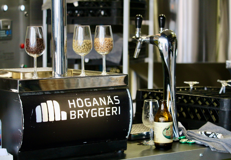 En bild som visar en del av en bryggerianläggning med en bardisk. På disken står tre vinglas fyllda med olika spannmål, sannolikt för bryggning. En öltappkran står till höger, bredvid en tom ölflaska och ett tomt vinglas. En skylt med texten "HÖGANÄS