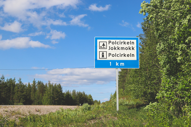 En blå vägskylt med vit text står vid sidan av en grusväg, omgiven av grön vegetation och barrträd. På skylten står det "Polcirkeln Jokkmokk", "Polcirkeln" och distansen "1 km". En blå himmel med vita moln syns i bakgrunden.