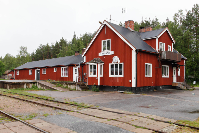 En stor, röd byggnad med vita fönster och en svart tak, sannolikt en gammal stationsbyggnad. Framför byggnaden ligger järnvägsspår och en asfalterad yta. I bakgrunden syns grön skog och en grå sky utan tydliga soltecken.