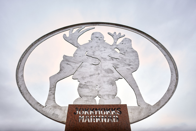 En metallskulptur föreställande en mänsklig figur och två renar, placerad under en gråmulen himmel. Skulpturen är inkapslad i en oval ring och har texten "JOKKMOKKS MARKNAD" utskuren i basen av rostfärgad metall. Den mänskliga figuren bär traditionel