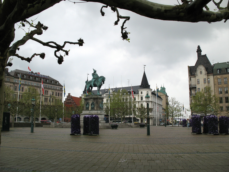 En stor öppen plats med kullersten kantas av byggnader och träd. I mitten står en ryttarstaty. Några flaggor syns framför en byggnad med 'Scandic Hotel' på taket. Himmelen är mulen. Lövträdens grenar syns i förgrunden