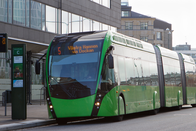 En modern grön ledbuss står parkerad vid en busshållplats. Bussen har destinationsskyltar som visar linje '5 Västra Hamnen via Dockan'. I bakgrunden syns moderna byggnader med stora glasfönster som reflekterar omgivningen.