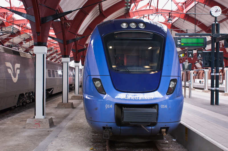 En blå Skånetrafiken-tåg står redo för avgång på en tågplattform under ett rött valvformat tak inomhus. Till vänster syns ett annat tåg bredvid vita pelare. En klocka och en digital informationstavla är synliga i bakgrunden till höger.