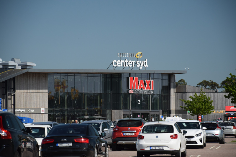 En bild som visar en shoppinggalleria med texten "GALLERIA center syd" och "MAXI STORMAMRNAD" på fasaden. Framför byggnaden finns en stor parkeringsplats fylld med flera personbilar. Himlen ovanför är klarblå. Träd och buskar syns i bakgrunden.