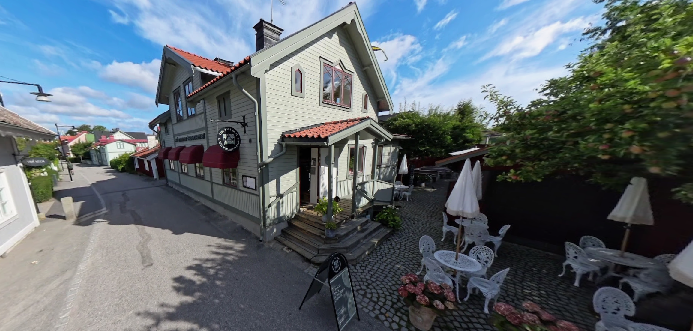 Bilden visar en ljusgrön träbyggnad med rött tak, som troligen är en restaurang eller ett café, med markiser över fönsterna och en uteservering med vita möbler. Till vänster syns en gata kantad av andra hus och grön vegetation; himlen är blå med vita