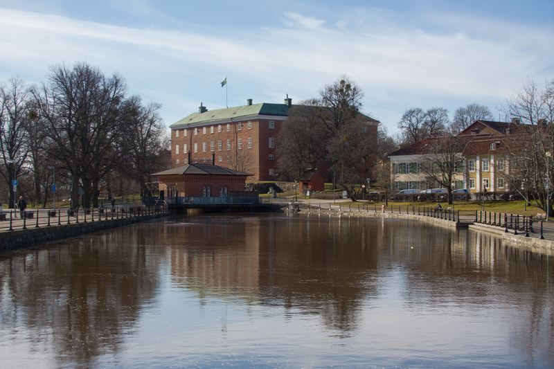 En bild som visar en flod eller kanal med byggnader längs stranden. Till vänster syns ett stort tegelhus med en svensk flagga på taket. Mellan vattnet och det stora tegelhuset finns en mindre tegelbyggnad. Till höger finns ytterligare byggnader och t
