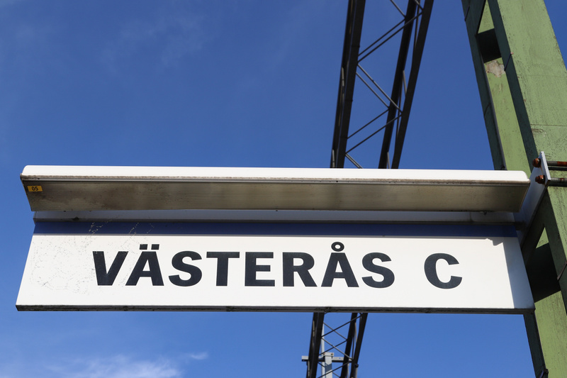 En skylt med texten "VÄSTERÅS C" i svarta bokstäver på vit bakgrund, monterad under en takliknande struktur. På skyltens övre vänstra del syns en gul etikett med texten "05". Bakgrunden består av en klarblå himmel med tunna vita moln. En grön stolpe