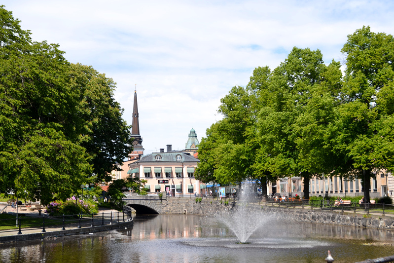 En bild som visar en stadsmiljö med en kanal i förgrunden där en fontän sprutar vatten. Längs kanalens sidor växer stora gröna träd och i bakgrunden syns flera byggnader. Ett kyrktorn med en spira sträcker sig mot den ljusa himlen.