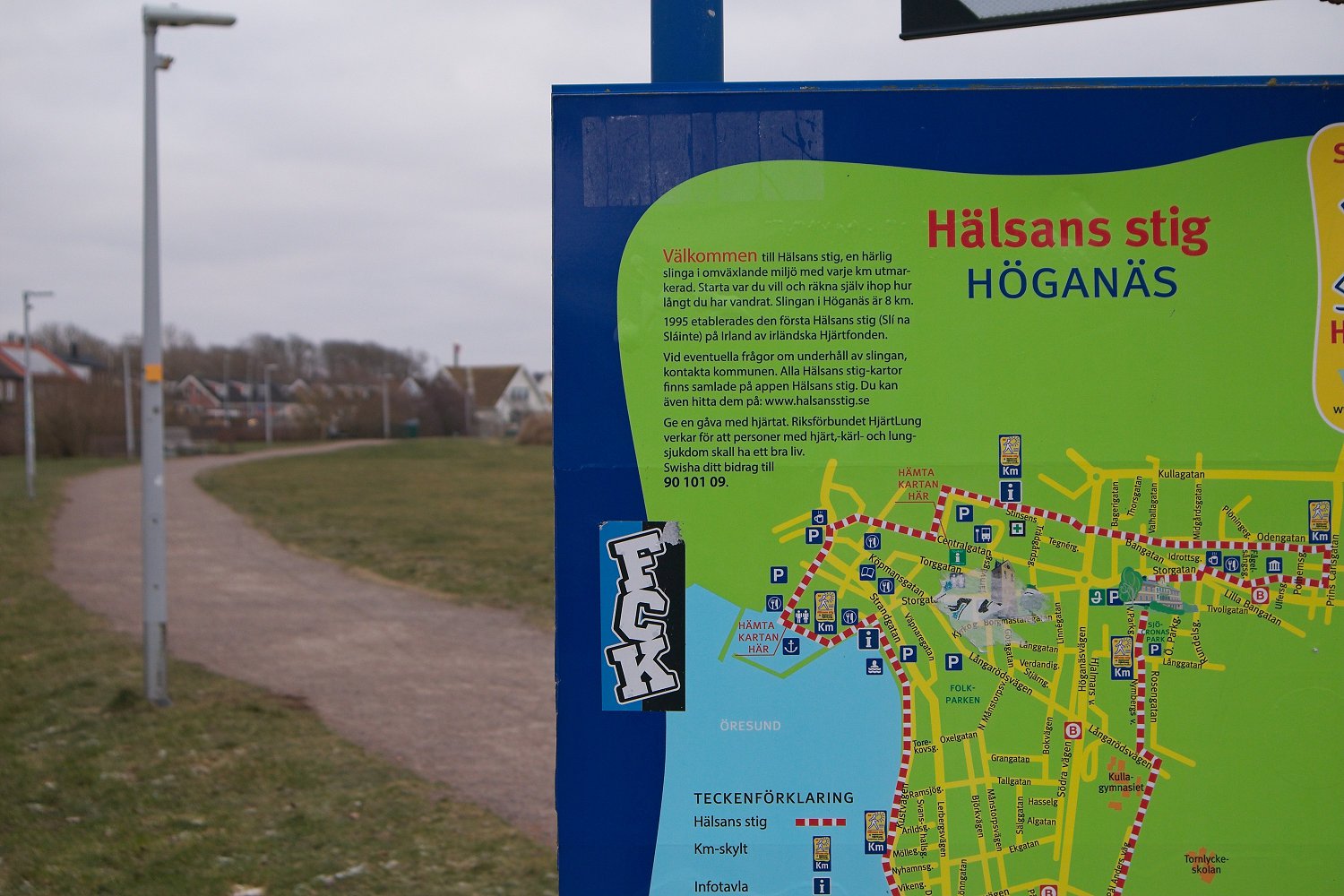 En blå informationstavla till höger i bilden med texten ”Hälsans stig HÖGANÄS” och en översiktskarta över området runt Öresund. På vänster sida syns en grusväg intill en gräsmatta, ett flertal belysningsstolpar och hus i bakgrunden. En klisterlapp si