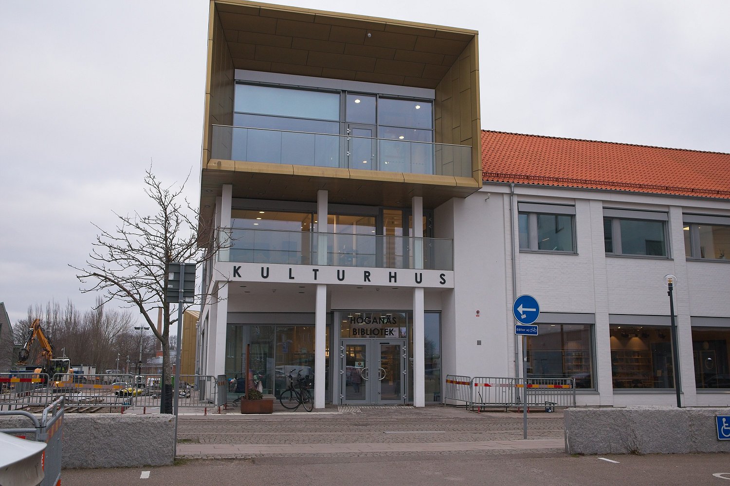 En fasad på ett "KULTURHUS" med texten "HÖGANÄS BIBLIOTEK" ovanför entrén. Byggnaden har vita väggar, stora fönsterpartier, en modern överbyggnad i guldfärg, och ett tegeltak. Utanför finns en cykel, ett träd och byggarbetsplatsutrustning i bakgrunde